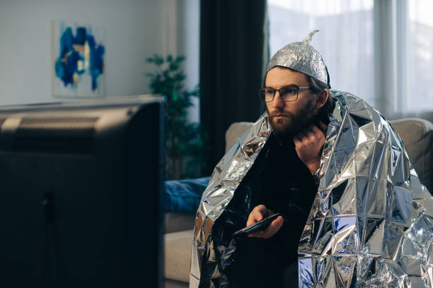 A young man wearing a tinfoil hat and wrapped in a tinfoil blanket is sitting on the couch in front of the TV with a remote control in his hands. A young man wearing a tinfoil hat and wrapped in a tinfoil blanket is sitting on the couch in front of the TV with a remote control in his hands. tinfoil barb barbonymus schwanenfeldii stock pictures, royalty-free photos & images