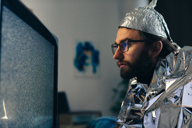 Conspiracy Theory on Screen: Man in Tin Foil Hat and Blanket Observing TV Interference Conspiracy Theory on Screen: Man in Tin Foil Hat and Blanket Observing TV Interference tinfoil barb barbonymus schwanenfeldii stock pictures, royalty-free photos & images