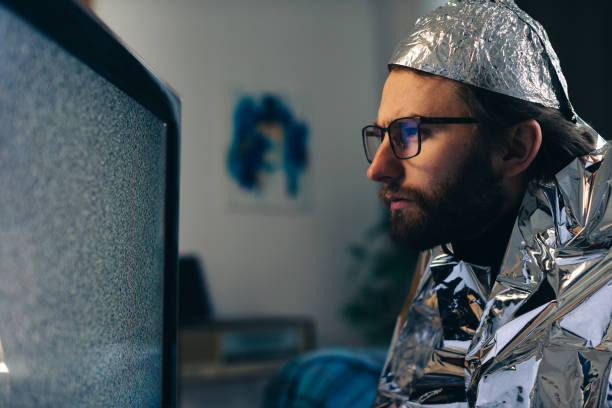 Conspiracy Theory on Screen: Man in Tin Foil Hat and Blanket Observing TV Interference Conspiracy Theory on Screen: Man in Tin Foil Hat and Blanket Observing TV Interference tinfoil barb barbonymus schwanenfeldii stock pictures, royalty-free photos & images
