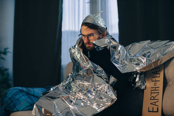 A man in a tinfoil hat is wrapped in a tinfoil blanket on the sofa at home. A man in a tinfoil hat is wrapped in a tinfoil blanket on the sofa at home. tinfoil barb barbonymus schwanenfeldii stock pictures, royalty-free photos & images