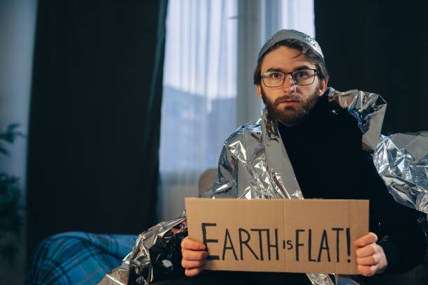 Flat Earth Advocate: Man in Tin Foil Hat and Blanket Holds Sign 'Earth is Flat Flat Earth Advocate: Man in Tin Foil Hat and Blanket Holds Sign 'Earth is Flat tinfoil barb barbonymus schwanenfeldii stock pictures, royalty-free photos & images