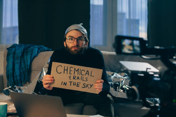 A conspiracy theorist shoots pseudoscientific videos on camera. A man in a tinfoil hat and a sign in his hands sits on a couch in front of the camera. A conspiracy theorist shoots pseudoscientific videos on camera. A man in a tinfoil hat and a sign in his hands sits on a couch in front of the camera. tinfoil barb barbonymus schwanenfeldii stock pictures, royalty-free photos & images
