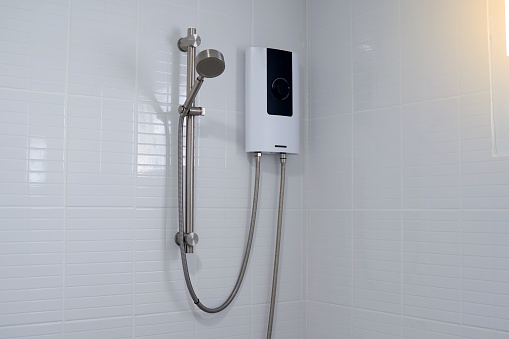 Water heater in black and white tones and a metal hand shower set mounted on a white bathroom wall.