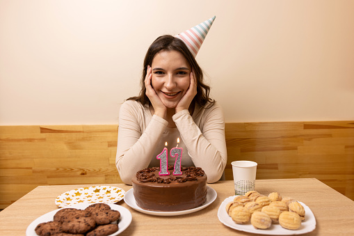 A girl sits in front of a table with a festive cake, in which a candle is lit in the form of the number 17. The concept of a birthday celebration.