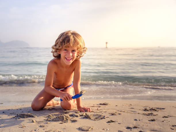 Smiling boy playing with toy shovel on sandy beach Charming curly haired boy playing with plastic shovel on wet sandy beach washed by clear sea and looking at camera with happy smile child no top at beach stock pictures, royalty-free photos & images