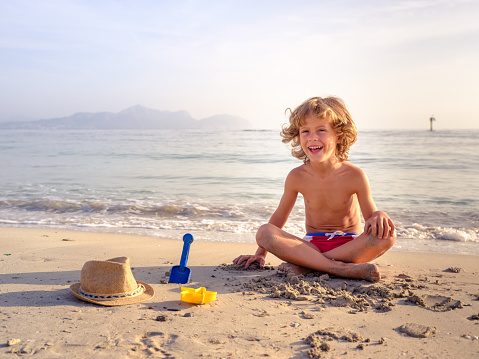 Little happy boy in summer hat playing with sand on the tropical beach, looking at the camnera with big smile on his face. Beautiful landscape. Blue sky and sea water background. Kid on vacation.