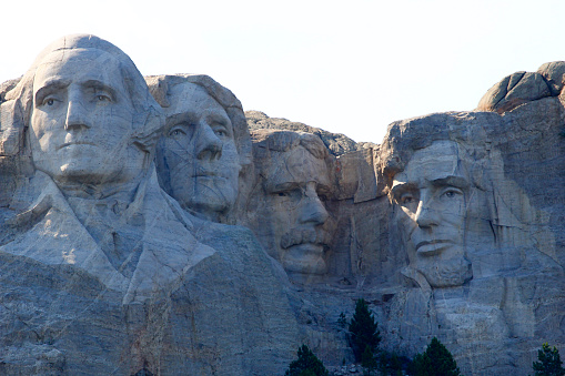Front view of Mount Rushmore. The carved faces of the four historical figures, George Washington, Thomas Jefferson, Theodore Roosevelt and Abraham Lincoln are framed by a brilliant stormy sky and tree branches. Bright green coniferous trees in the foreground provide contrast to the granite stone.