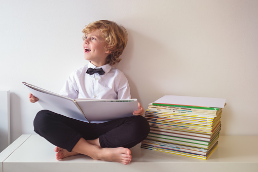 Full body of cheerful boy in formal wear looking into distance with smile while reading book on white background near heap of textbooks