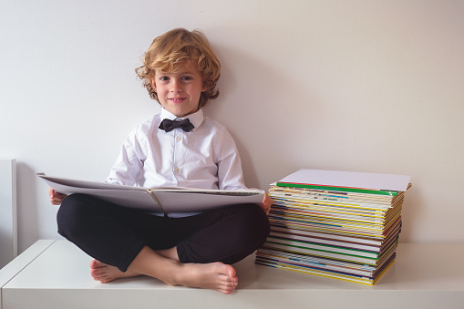 Full body of cheerful boy in formal wear looking at camera while reading textbook near pile of literature on white background