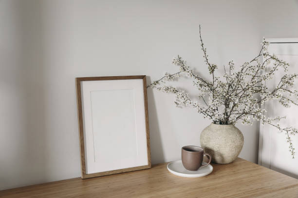 istock Vertical wooden picture frame, poster mockup in sunlight. Spring, easter composition. Eleganrnterior, home office still life. Blooming cherry plum tree branches in vase. Wooden table. Lateral view. 2112847229