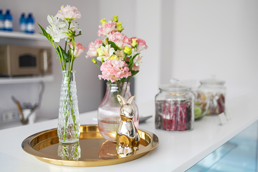 A clear glass vase with delicate white and pink flowers alongside a shiny golden rabbit figurine on a reflective tray, set against a modern kitchen backdrop.