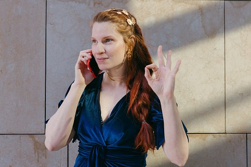 Confident woman in a luxurious blue dress speaking on the phone and gesturing 'okay', displaying assurance and elegance.