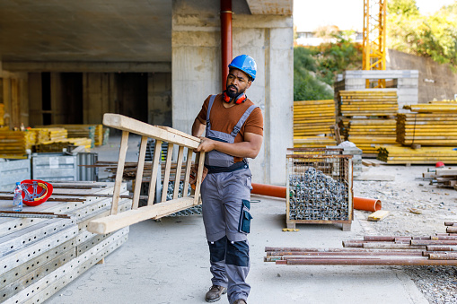 A Young African-American Building Worker with Protective Helmet is Working at the Building Site and Carrying Ladders.