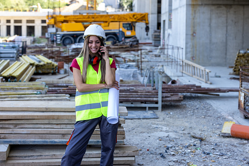 Young Female Building Worker is Standing at a Construction Site and Communicate with Coworkers or Friends Using a Smart Phone.