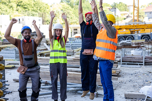 Photo of Multi-Ethnic Group of Building Workers with Protective Helmets who is Feeling Successful at the Construction Site and Celebrating with Raised Arms.