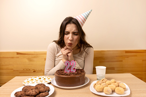 A girl sits in front of a table with a festive cake and blows out a candle in the form of the number 22. The concept of a birthday celebration.