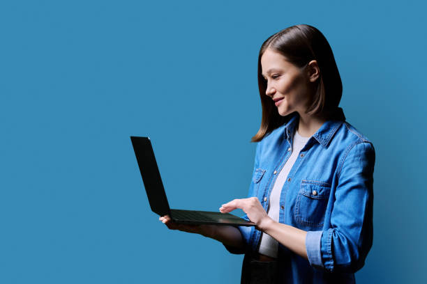 istock Young smiling woman using laptop, profile view on blue background 2112913793