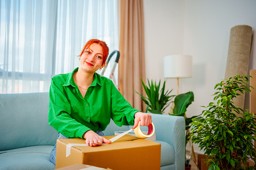 Young woman  uses adhesive tape to packing cardboard box. Moving home concept, looking at camera