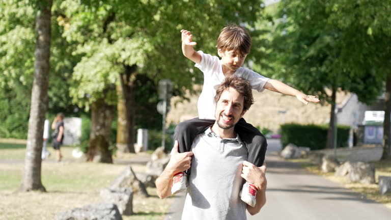 Under Bright Sun, Affectionate Father Enthusiastically Carries Son On Shoulder During Lush Park Walk
