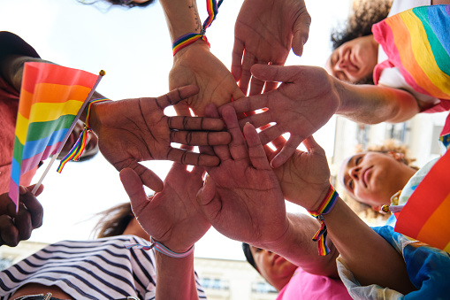 A group of multiracial people with rainbow LGBTQ bracelets stacking hands. Scene is one of unity and support for the LGBTQ community.
