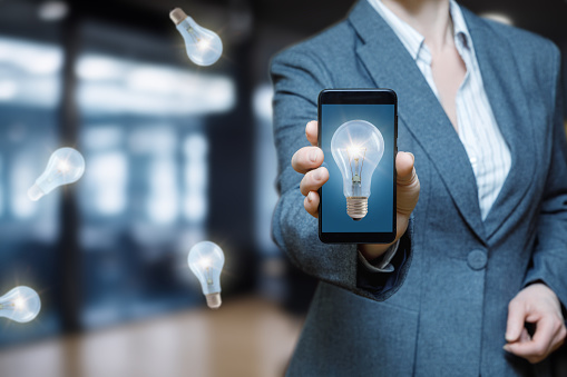 A businesswoman shows a cell phone with a light bulb burning on the display. The concept of an idea in a cell phone.