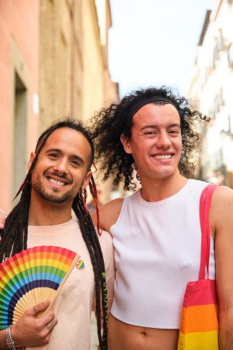 Gay couple smiling, looking at camera and holding a rainbow fan during International Pride Day.