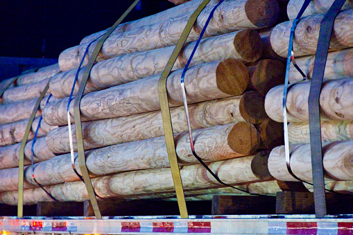 Heavy straps and metal bands hold a load of finished round lumber onto a semi-truck trailer.