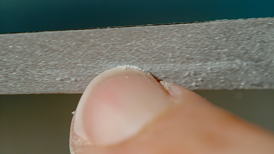 MACRO, DOF: Detailed closeup shot of a young woman filing her nails with a rough nail. Unrecognizable white female's fingernail gets filed, sending smooth particles of filed nail flying in the air.