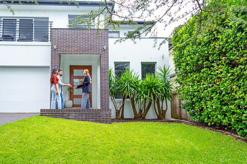 Real estate agent showing a couple a new house. The house is contemporary. All are happy and smiling and shaking hands. The couple are casually dressed and the agent is in a suit. The house has a front lawn and a brick facade