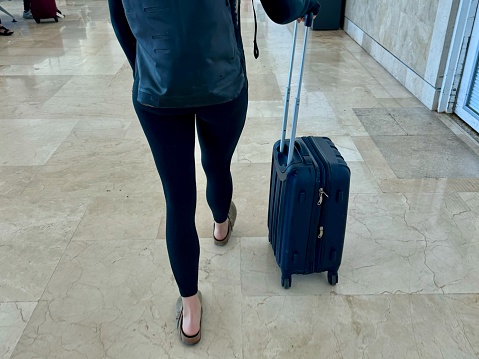 Back view of a woman's back and legs wearing black leggings and slip-on shoes, carrying a black backpack and wheeling a small, hard-shell, carry-on suitcase through the Cancun airport
