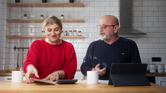 Senior parents sit in kitchen doing paperwork, checking calculating bills bank loan payment, discuss unpaid debt taxes. Old grandparents family money problem concept