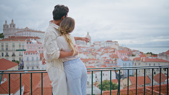 Embracing lovers contemplating seascape together at mediterranean town vacation back view. Tanned enamoured man cuddling loving woman at romantic date. Relaxed couple admiring houses in tender hugs