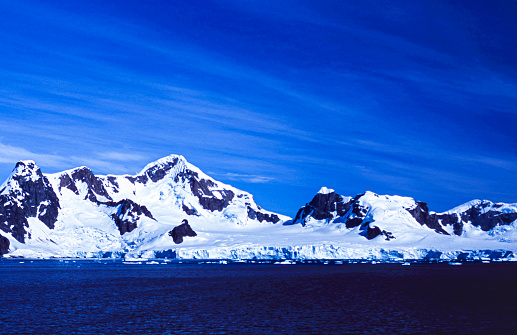 Snow covered mountain peaks and glacier formations, in an Antarctica bay, which contains small floating icebergs.\n\nTaken in Antarctica.
