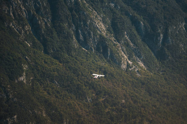 A small airplane is captured in flight against the backdrop of steep, wooded mountains. This aerial view highlights the contrast between the man-made aircraft and the natural rugged terrain below.