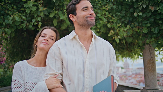 Happy newlyweds resting park together closeup. Hispanic tanned man holding book contemplating cityscape with loving girlfriend. Satisfied smiling sweethearts calm romantic date at mediterranean city