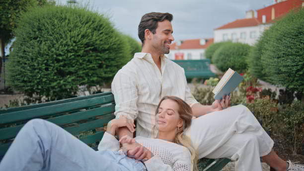 Relaxed newlyweds enjoying novel park zoom out. Man reading book for girlfriend Relaxed newlyweds enjoying novel at town park zoom out. Happy smiling couple embracing at nature square relax. Tender man reading book for girlfriend. Carefree sweethearts spending romantic date city zoom out stock pictures, royalty-free photos & images