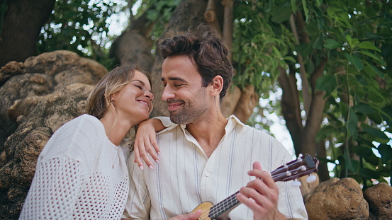 Enamoured newlyweds enjoying guitar music at woods closeup. Handsome macho playing ukulele for loving woman. Young lady kissing boyfriend chick at park. Love couple dating at forest. Happiness concept
