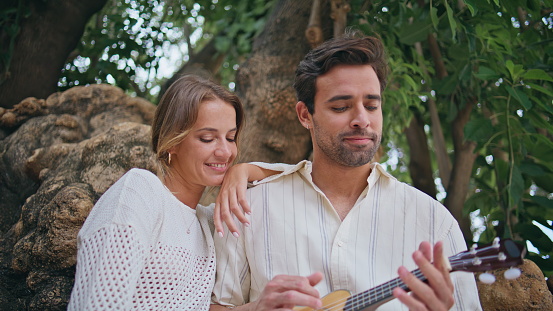 Tender lovers relaxing guitar music at greenery nature closeup. Loving man woman enjoying song at big tree park. Handsome unshaven guy holding ukulele relaxing with girlfriend. Love pair romantic date