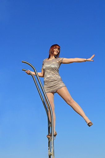A young woman in a dress stands high up on a ladder and stretches out one arm and one leg