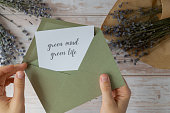 Female hands taking paper card note with text GREEN MIND GREEN LIFE from envelope. Lavender flower. Top view, flat lay. Concept of eco sustainability lifestyle mental spiritual health