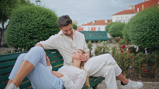 Enamoured man caressing woman face at cozy park bench zoom on. Beautiful woman looking at boyfriend feeling happy at weekend. Loving couple holding hands enjoying romantic honeymoon date together