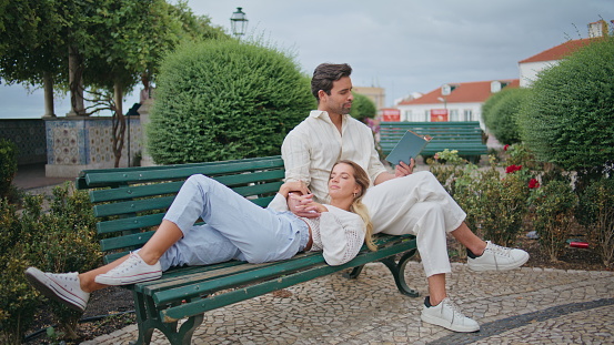 Tender newlyweds resting bench together at park. Hispanic tanned man reading book aloud relaxing with loving girlfriend at greenery city nature. Happy sweethearts calm romantic date at bushes place