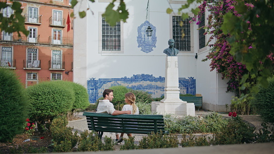 Love couple sitting bench talking at urban park. Affectionate man embracing girlfriend enjoying time together at romantic date. Lovely pair bonding at greenery nature. Honeymoon vacation concept