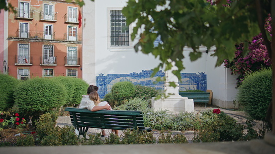 Romantic pair embracing tenderly sitting park bench back view. Brunette man bonding to girlfriend at city date. Enamoured couple enjoying time together at greenery square vacation. Love concept