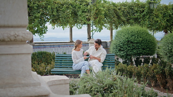Cheerful couple sitting bench laughing at urban park. Affectionate man holding book telling funny story to woman at romantic date. Lovely pair enjoying time together kissing at greenery nature