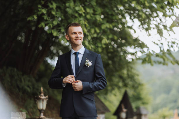 the groom in a black suit adjusts his jacket, poses against the background of a green tree. wedding portrait. - fashion model personal accessory suit tying ストックフォトと画像