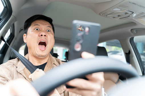 A man is driving a car and looking at his cell phone. He is surprised and excited. Concept of distraction while driving and the potential danger it poses.