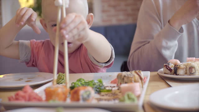 Cute boy eating sushi rolls with stick in sushi bar