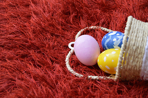 Colorful hand painted Yellow, blue and pink ornate Easter eggs in a wicker basket spilling out on bright vibrant red maroon carpet or rug background for of Easter holidays celebration with copy space. Apt for use as greeting cards, posters and backdrops.