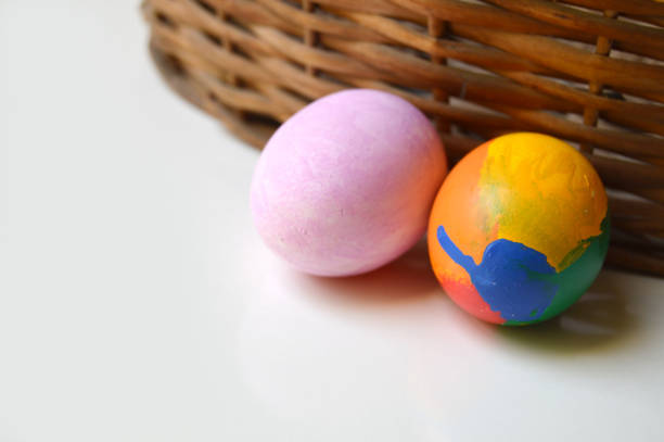 Two colorful hand painted pink and multi coloured green blue and yellow ornate Easter eggs on bright off white cream colored background against a wooden brown rustic woven wicker basket for of Easter holidays celebration with copyspace. Apt for use as greeting cards, posters wallpaper and backdrops.